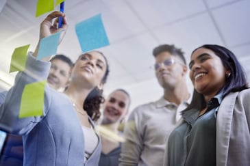 Grupo de colaboradores engajados colando e analisando notas adesivas em uma parede de vidro, ilustrando a coleta de feedback participativa durante uma pesquisa de clima organizacional.